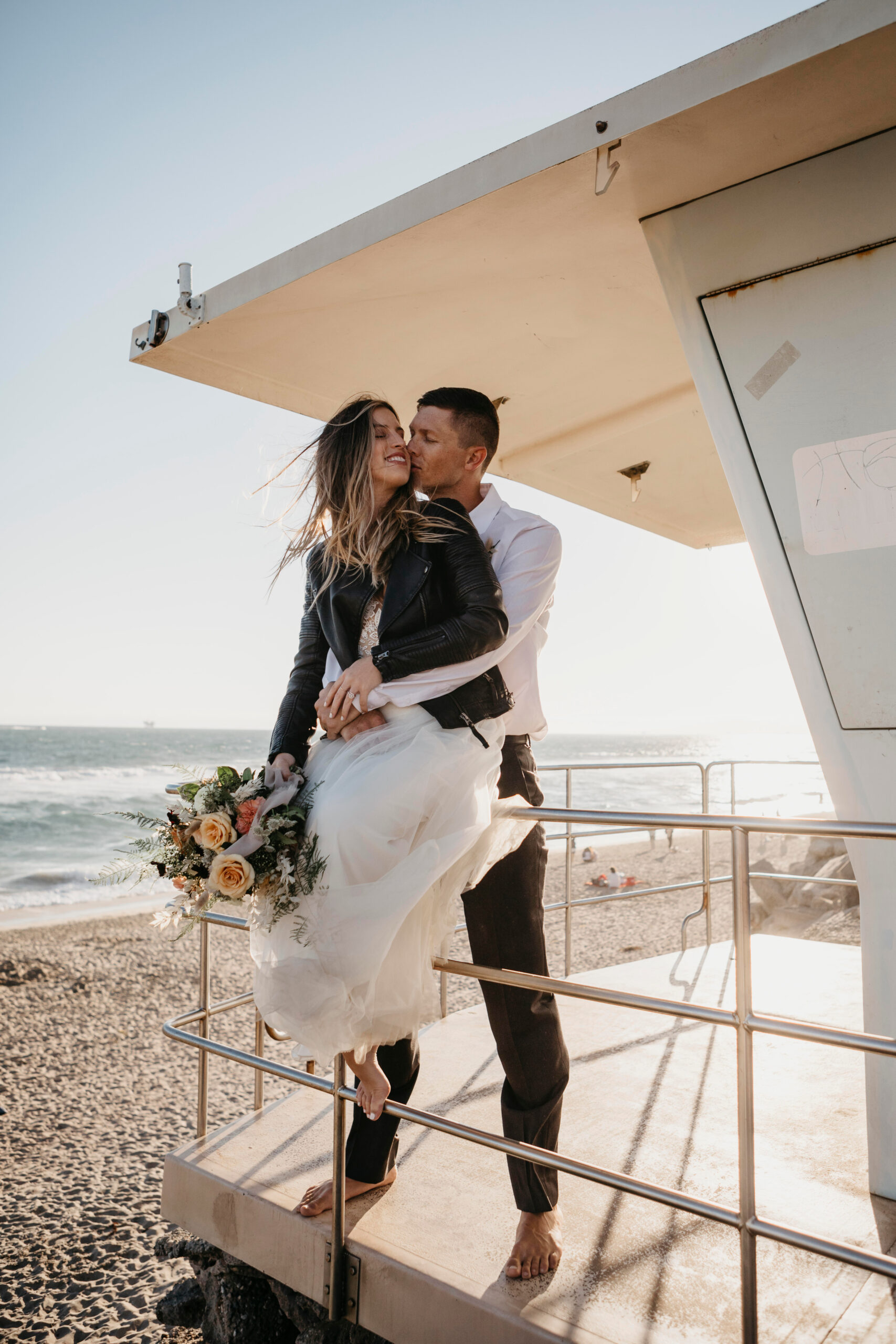 Noivos trocam um beijo em uma plataforma próxima à praia, com o oceano ao fundo. | Foto: Adobe Stock
