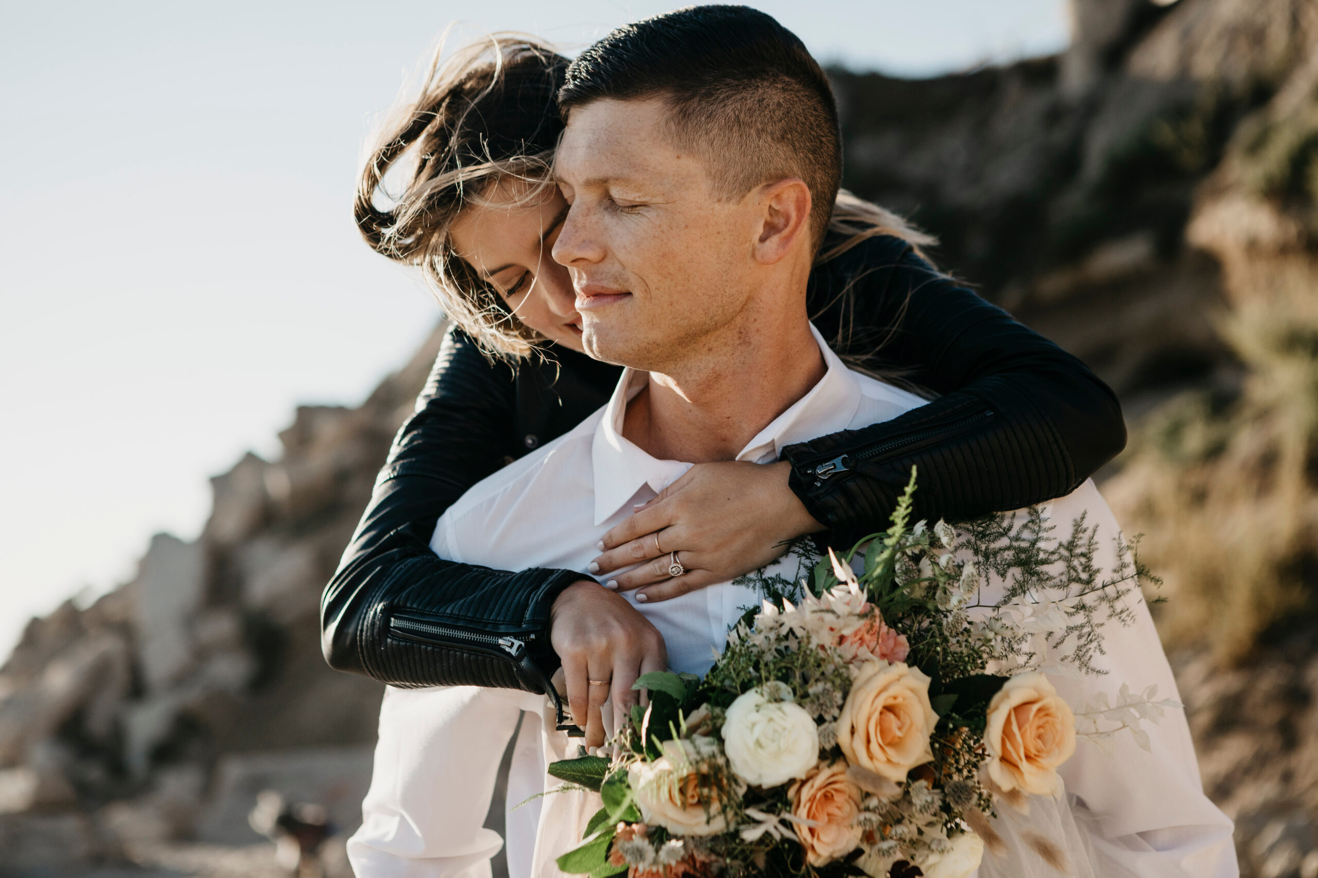 Noiva abraça o companheiro ao ar livre, segurando um buquê de flores em tons claros. | Foto: Adobe Stock