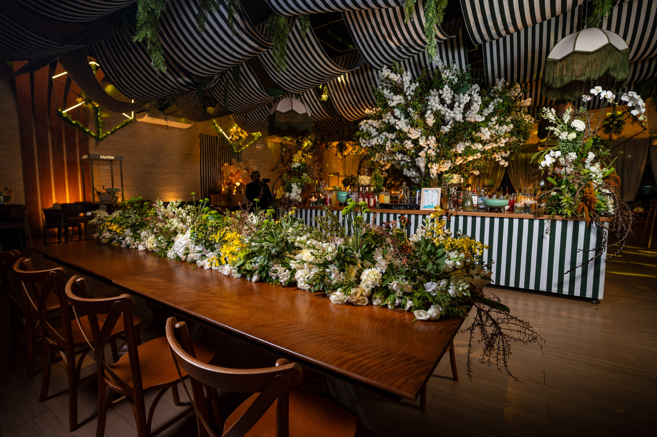 Mesa de madeira com arranjos florais em tons de branco e amarelo integra a decoração, marcada por tecidos listrados e elementos naturais. | Foto: Chris Machado