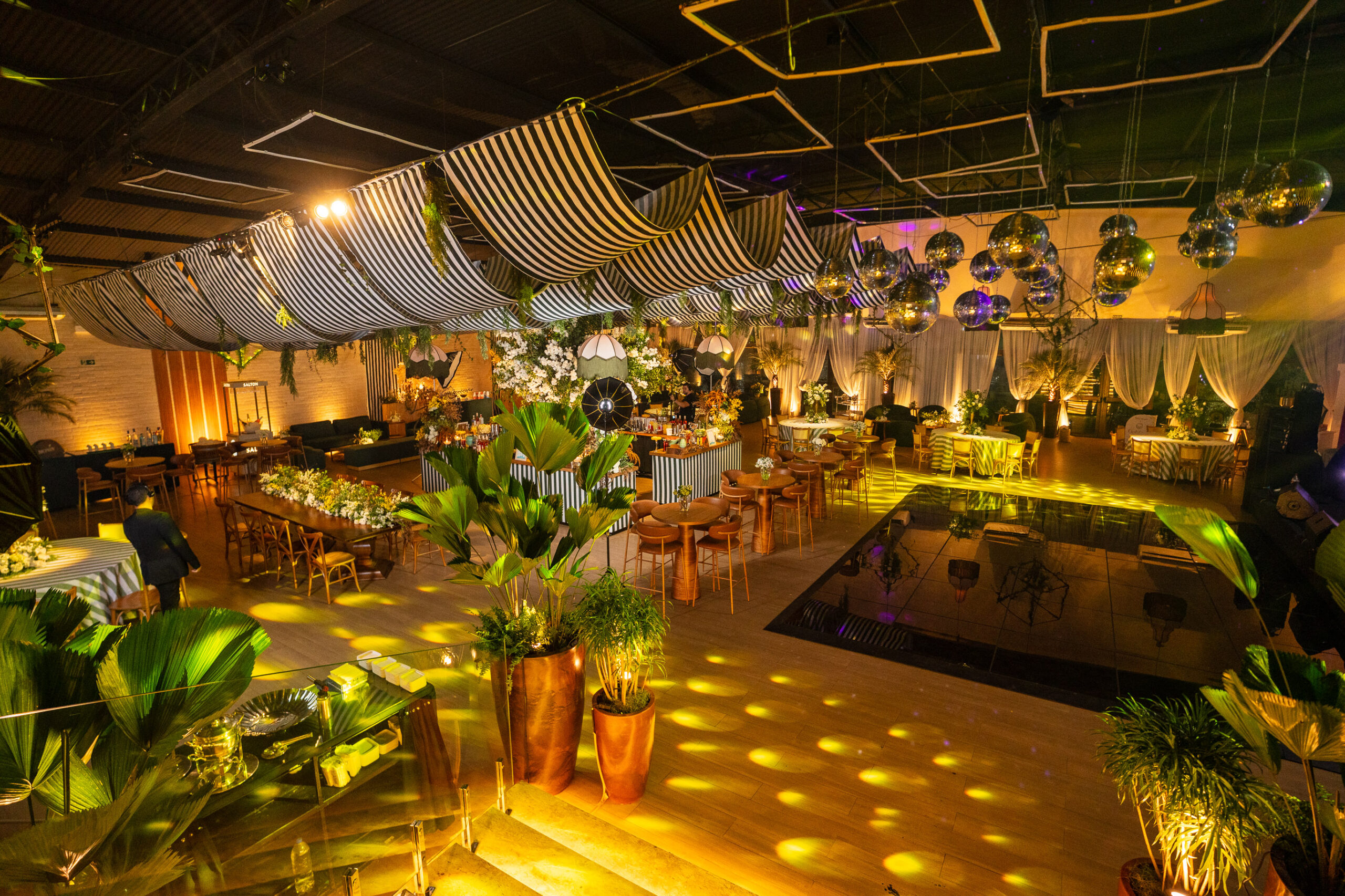Vista panorâmica da decoração do Ballroom Beach Club, com tecidos listrados, arranjos florais, plantas tropicais e globos espelhados sobre a pista de dança. | Foto: Chris Machado