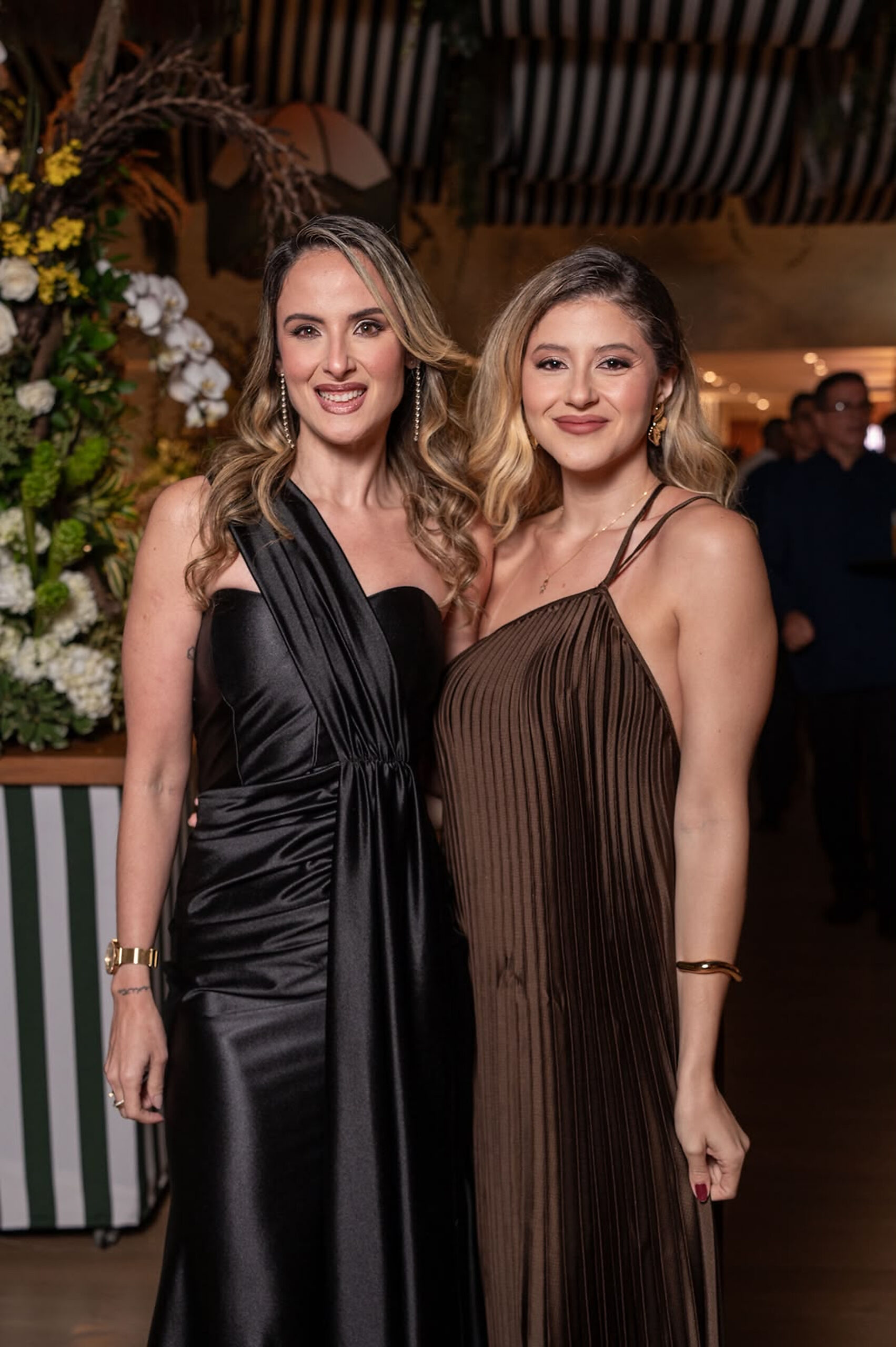 Camilla Martins e Gabriela Melo posam para fotografia ao lado de um arranjo de flores brancas durante a inauguração do Ballroom Beach Club. | Foto: Chris Machado