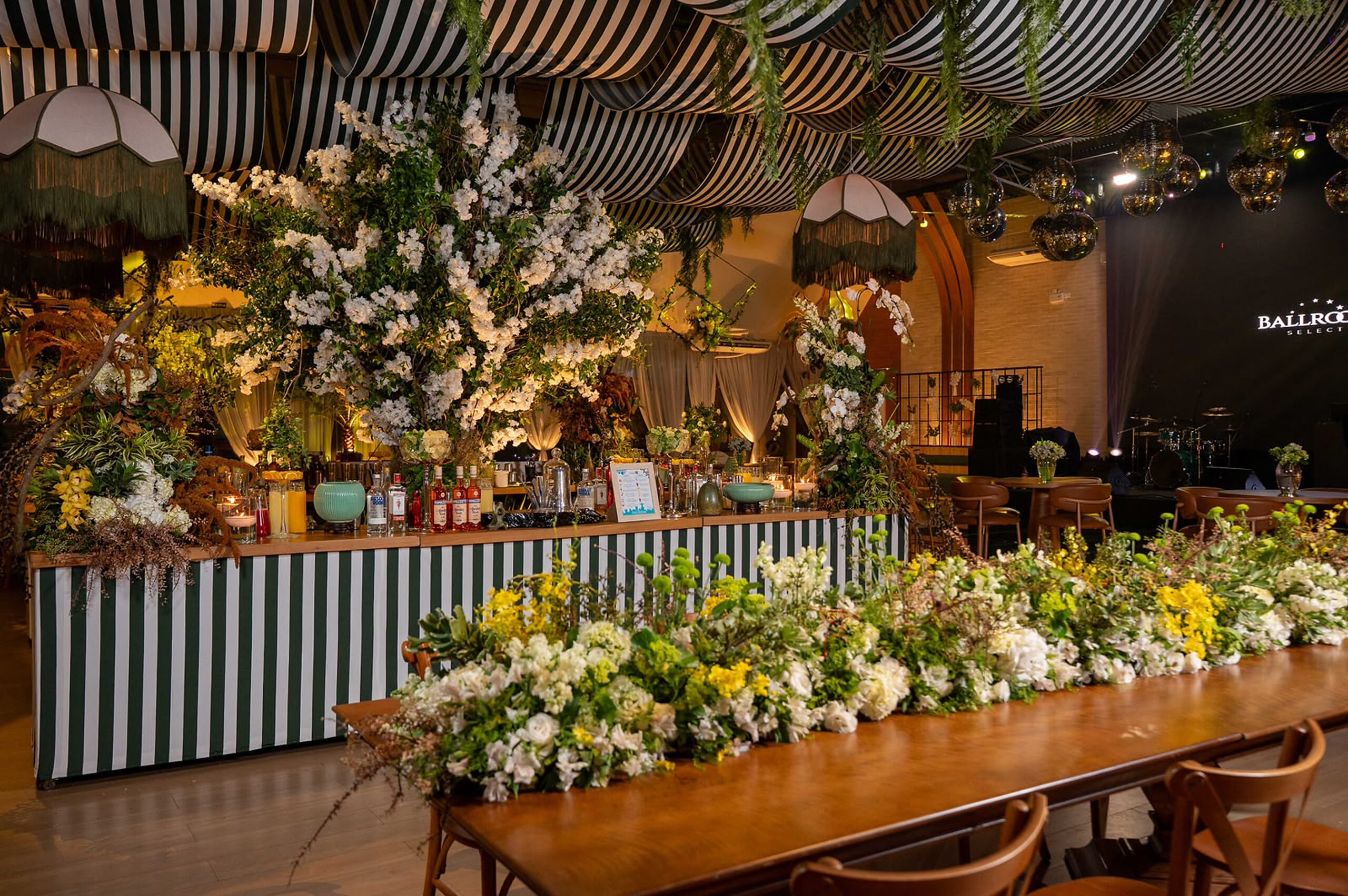 Mesa comprida com arranjo floral contínuo integra a ambientação sob tecidos listrados e luminárias pendentes. | Foto: Chris Machado