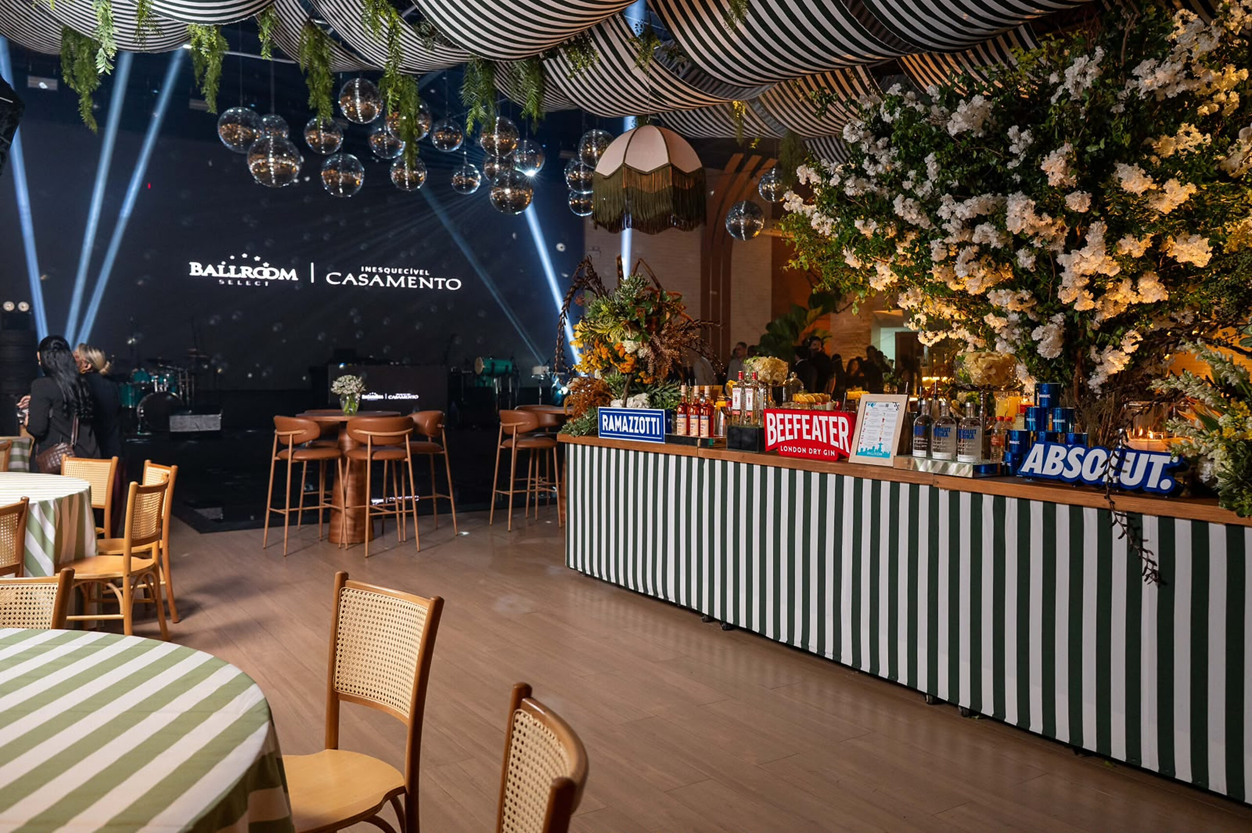 Balcão de bar com bebidas expostas e arranjos florais aparece ao lado do palco com as marcas Ballroom e Inesquecível Casamento. | Foto: Chris Machado