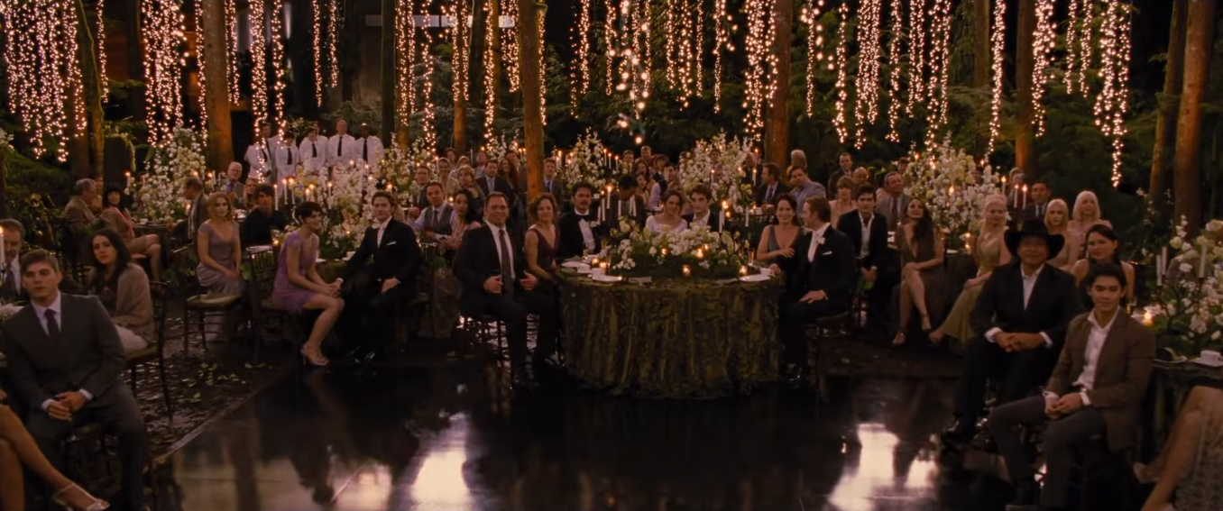O jantar de casamento de Bella e Edward acontece em meio à floresta, com mesas decoradas por arranjos florais brancos, velas e uma iluminação composta por centenas de pequenas luzes suspensas entre as árvores. | Reprodução/Paris Filmes