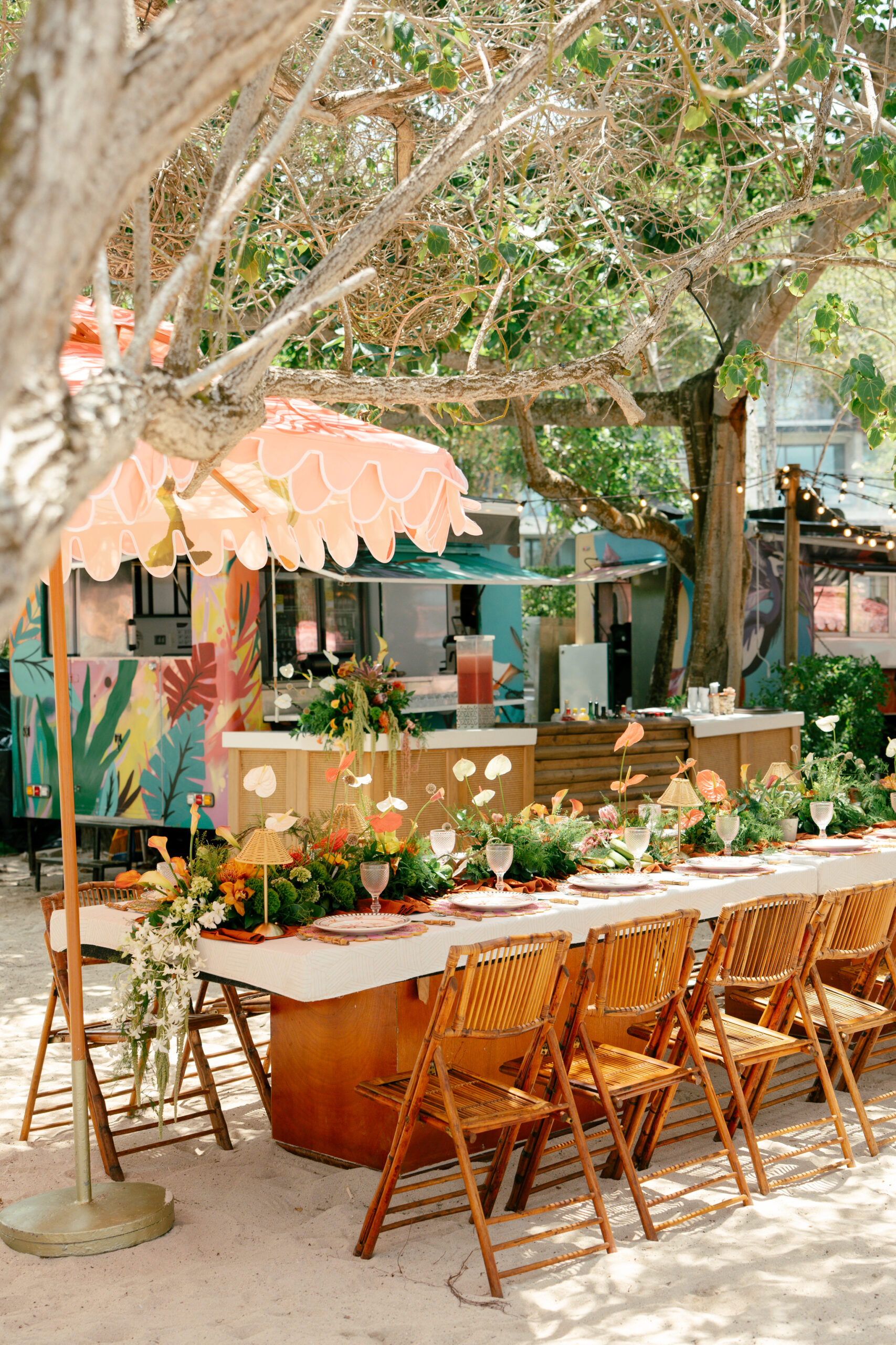 Mesas decoradas com flores tropicais e cadeiras de bambu compõem um espaço gastronômico sob as árvores, com estações de serviço ao fundo | Foto: Compañía Audiovisual