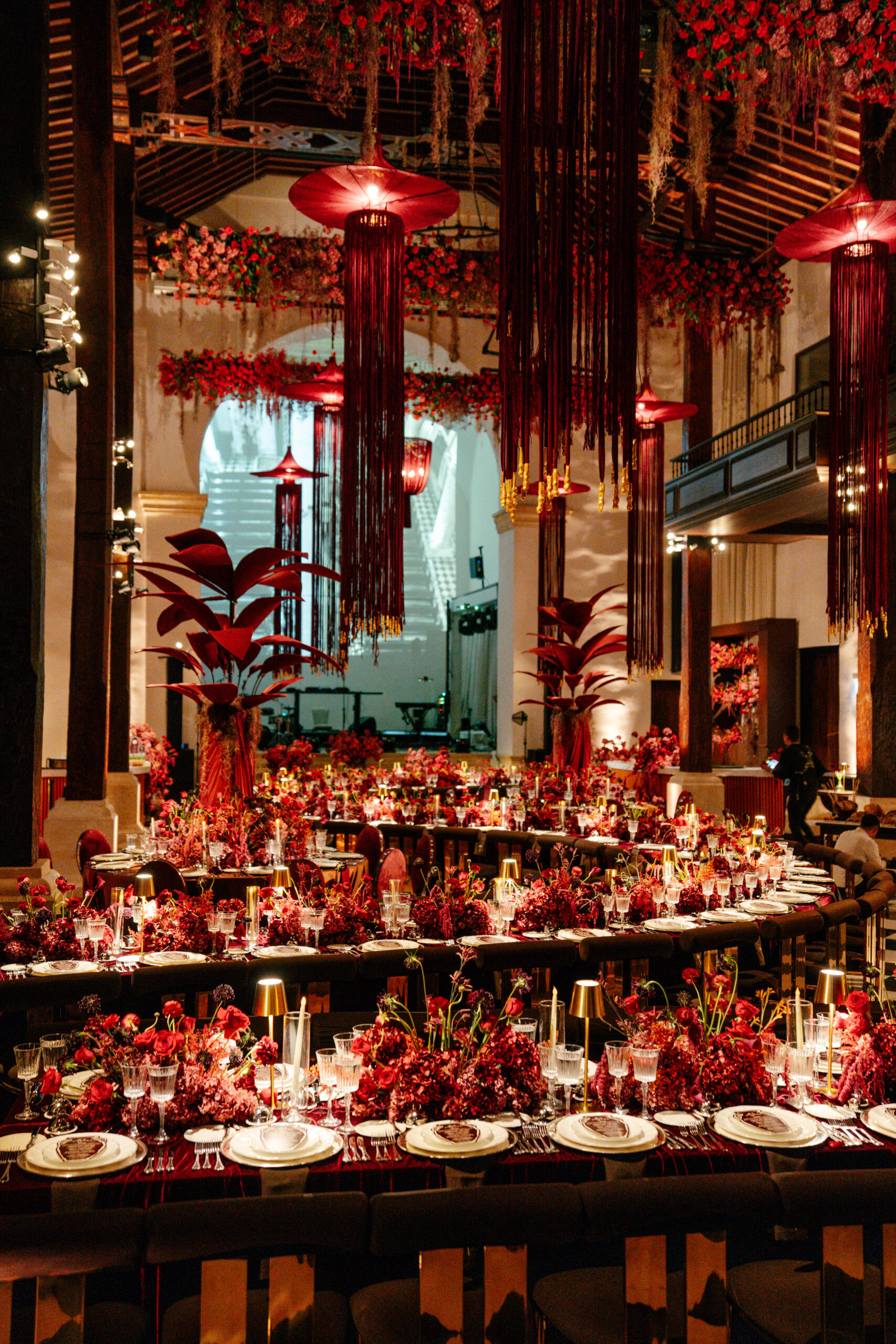 Composição floral em tons de vermelho e vinho, acompanhada de iluminação pontual e grandes estruturas suspensas, transforma o salão em um ambiente de celebração | Foto: Compañía Audiovisual