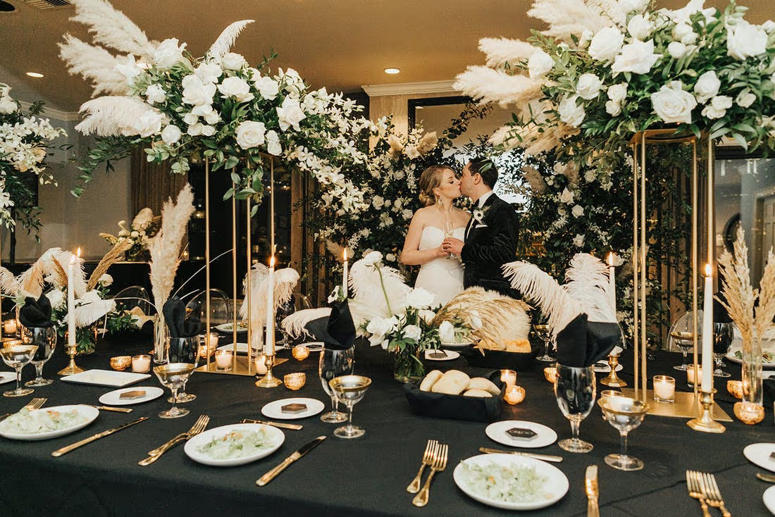 Recepção de casamento com decoração sofisticada inspirada na década de 1920, marcada por flores brancas, folhagens, plumas, velas e detalhes dourados, enquanto os noivos celebram ao centro do ambiente. I Foto: Party Slate