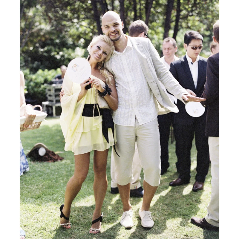 Casal de convidados posa durante um casamento ao ar livre, com ela usando vestido curto amarelo-claro, faixa preta na cintura e sandálias de salto, enquanto ele aposta em um conjunto de linho em tons claros, com blazer, camisa, bermuda e sapatos brancos, em uma composição descontraída para celebrações durante o dia. | Foto: Polly Alexandre Photography