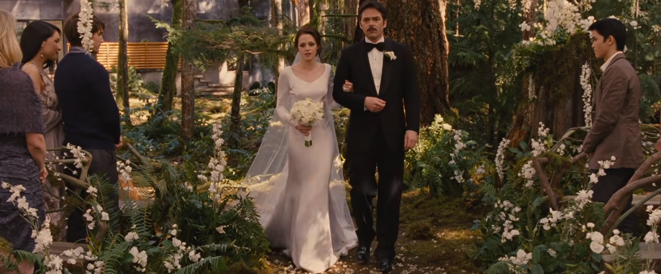 Bella caminha até o altar acompanhada pelo pai, Charlie Swan. O vestido de mangas compridas, o buquê branco e os arranjos florais ao longo do corredor compõem a estética romântica da cerimônia. | Reprodução/Paris Filmes