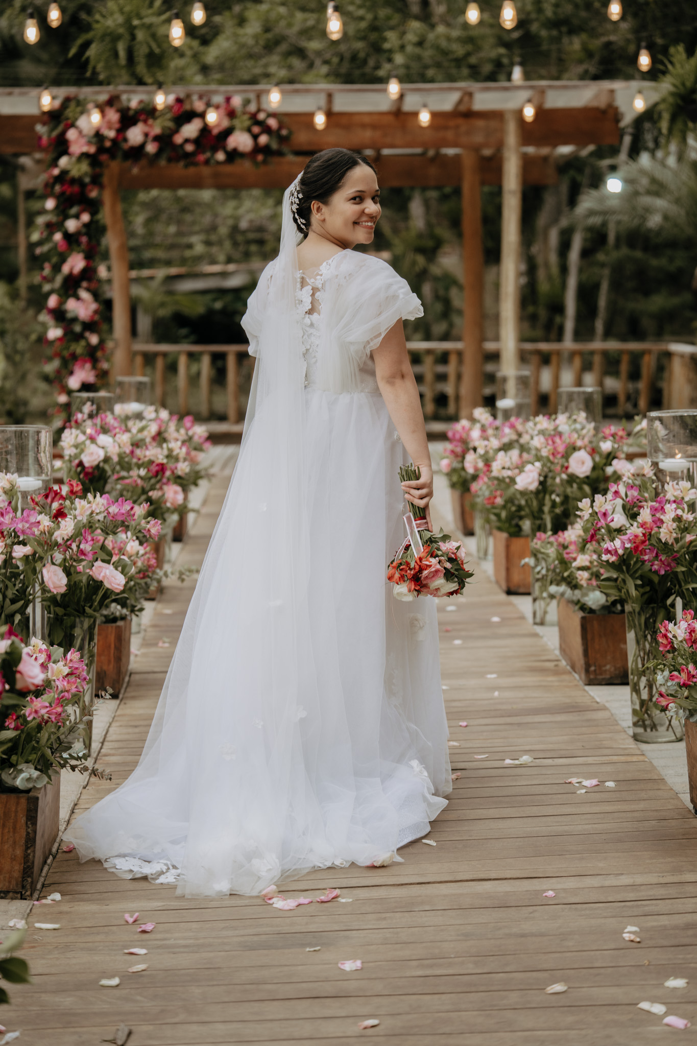 Noiva posa no corredor da cerimônia cercado por arranjos florais em tons rosados | Foto: Divulgação/Mi B'Antonni