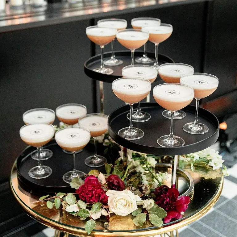 Uma sofisticada composição de taças de coquetéis, dispostas em bandejas pretas de diferentes alturas, integra a decoração do evento. Na base, um arranjo floral com rosas brancas, flores em tons de vinho e folhagens verdes acrescenta cor e delicadeza à composição. I Foto: The Knot
