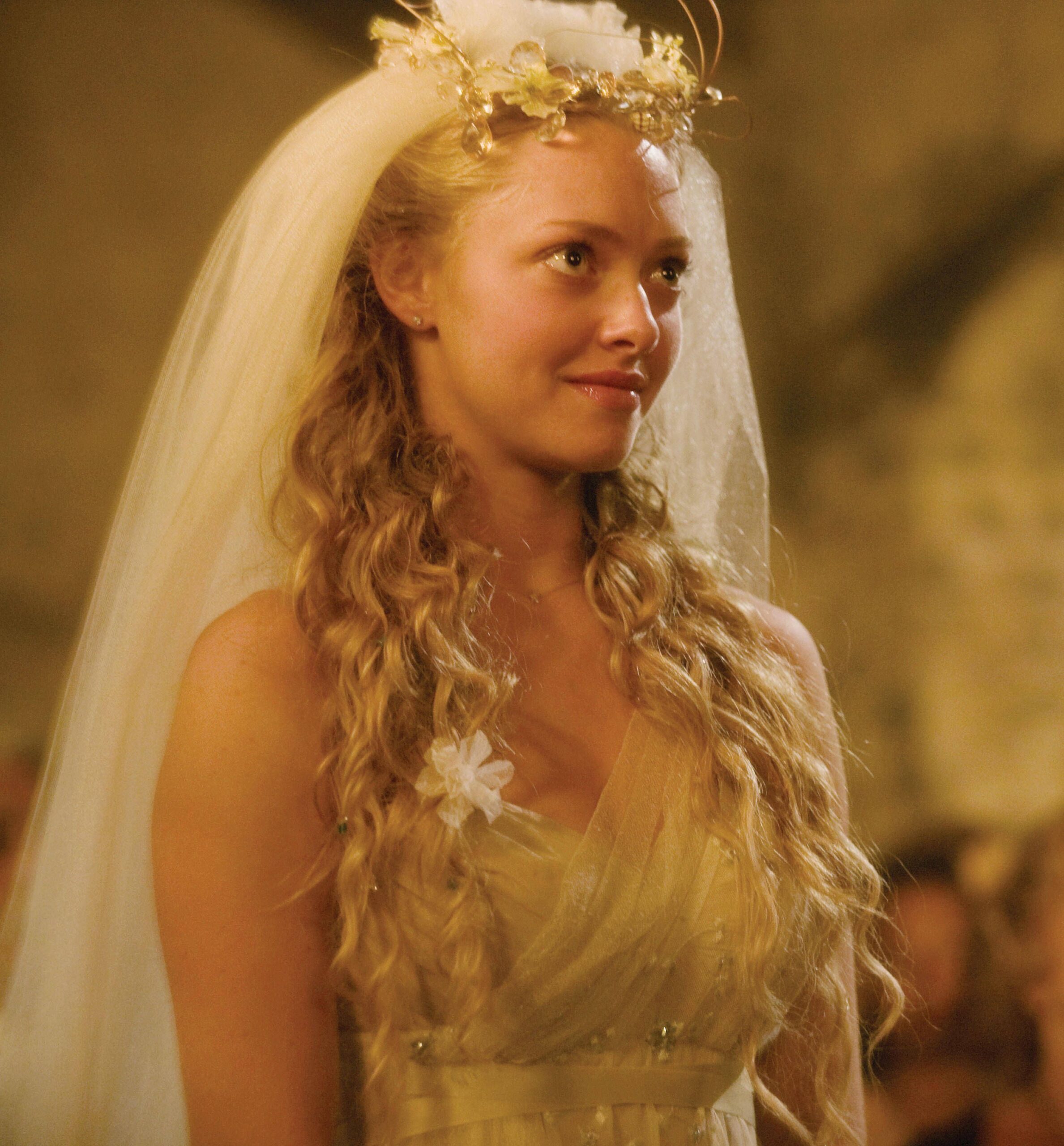 Sophie aparece com os cabelos soltos em ondas, uma coroa de flores e um longo véu. O vestido apresenta tecido leve, detalhes florais e uma modelagem de inspiração boêmia. | Reprodução/Universal Pictures