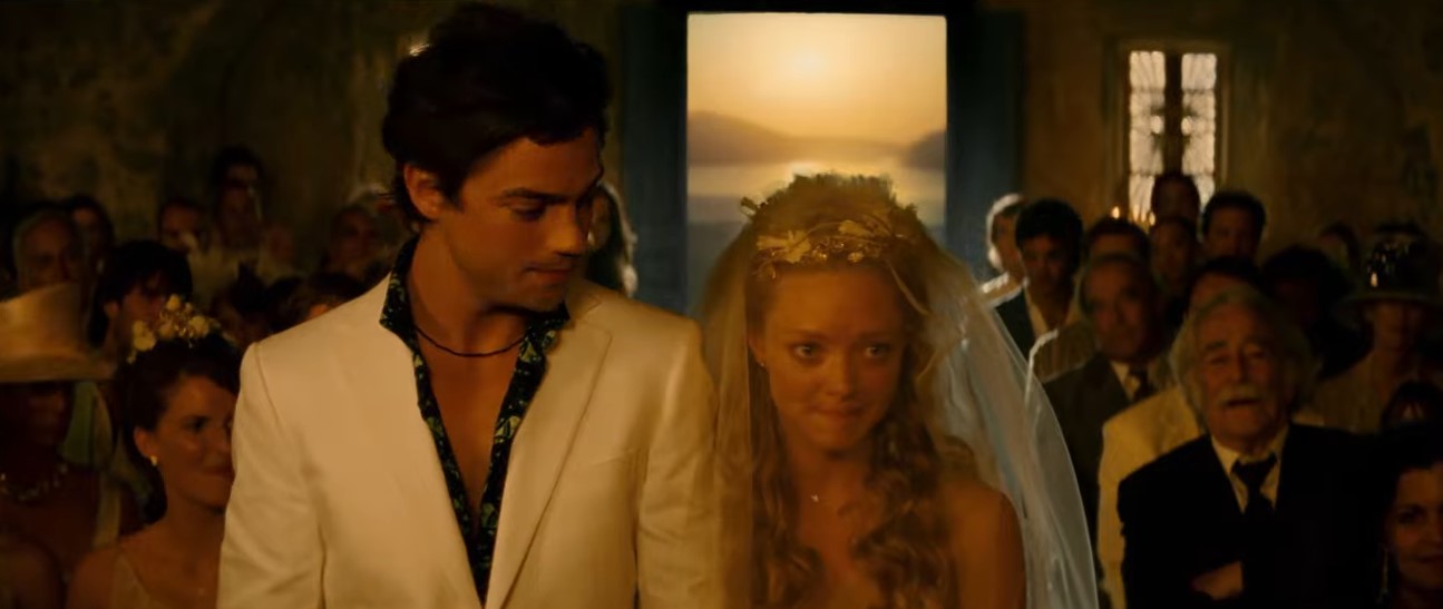 Sophie e Sky aparecem lado a lado durante a cerimônia, usando trajes leves que acompanham a atmosfera descontraída do casamento na ilha grega. | Reprodução/Universal Pictures