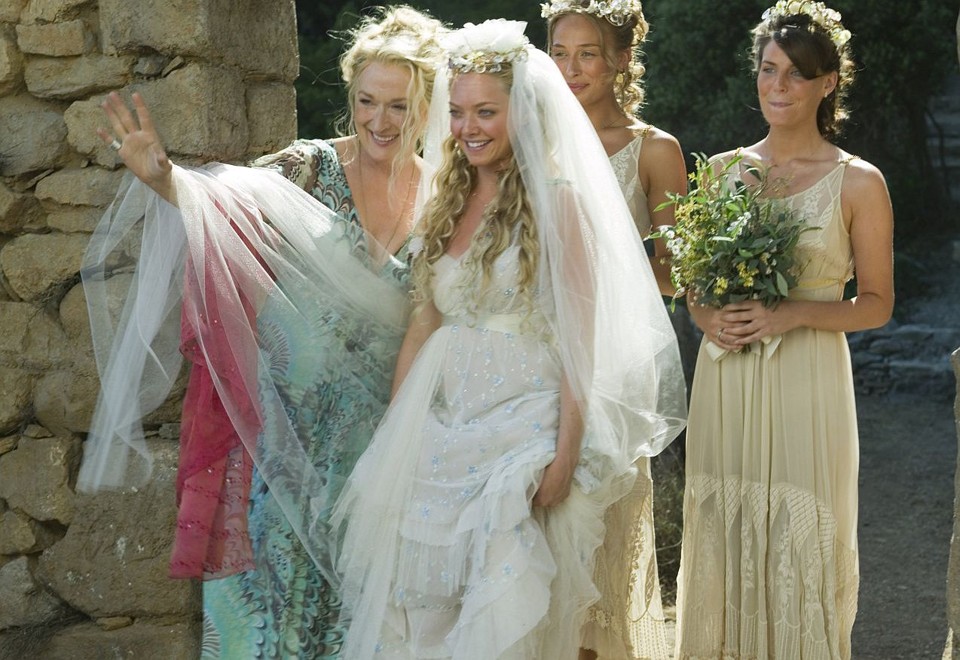 Sophie se prepara para o casamento ao lado de Donna e das amigas. O vestido apresenta aplicações delicadas, camadas de tecido fluido e um véu volumoso, complementado por uma coroa de flores. | Reprodução/Universal Pictures