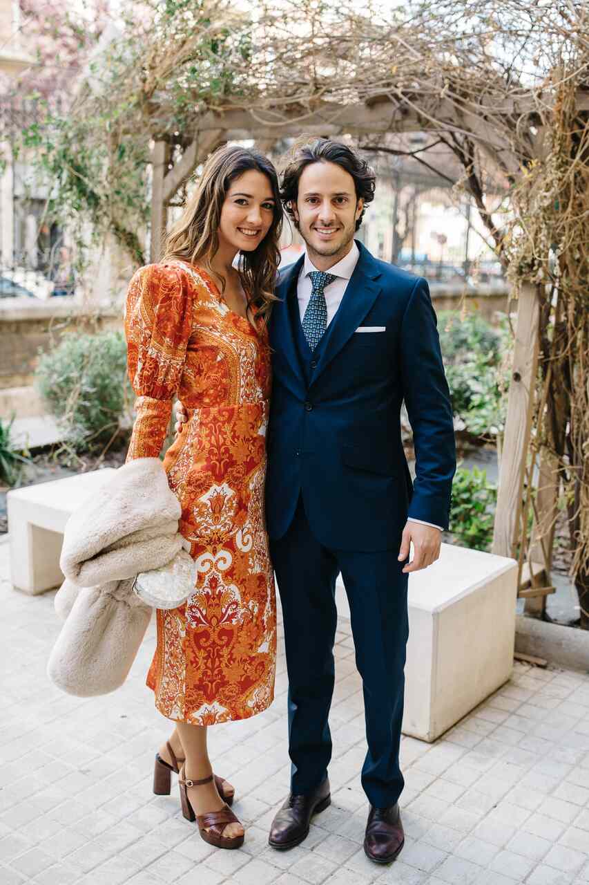 Casal posa em um jardim, com ela usando vestido midi estampado em tons de laranja, mangas longas e sandálias de salto, enquanto ele veste terno azul-marinho de três peças, camisa branca e gravata estampada, em uma composição elegante para um casamento durante o dia. | Foto: Divulgação / Lápis de Noiva