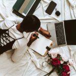 Mulher organiza anotações em uma agenda ao lado de um notebook, celular e flores, elementos que ilustram o planejamento de um casamento e o uso de ferramentas digitais. | Foto: Pexels / David Kwewum