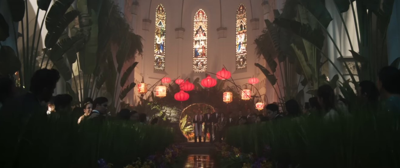 A cerimônia de casamento de Araminta e Colin transforma o interior de uma igreja em um jardim tropical, com grandes folhagens, flores coloridas, lanternas suspensas e iluminação cenográfica. | Reprodução/Warner Bros. Pictures