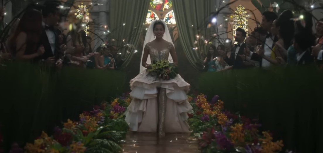 Araminta entra na cerimônia usando um vestido com aplicações brilhantes, saia estruturada e véu longo. O corredor, coberto por uma lâmina de água, reflete a iluminação e a decoração floral. | Reprodução/ Warner Bros. Pictures