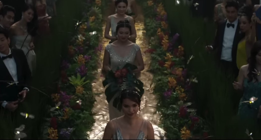 Madrinhas caminham pelo corredor da cerimônia, cercadas por arranjos tropicais e pontos de luz que remetem a vagalumes, em uma decoração marcada pela combinação de natureza e tecnologia. | Reprodução/Warner Bros. Pictures