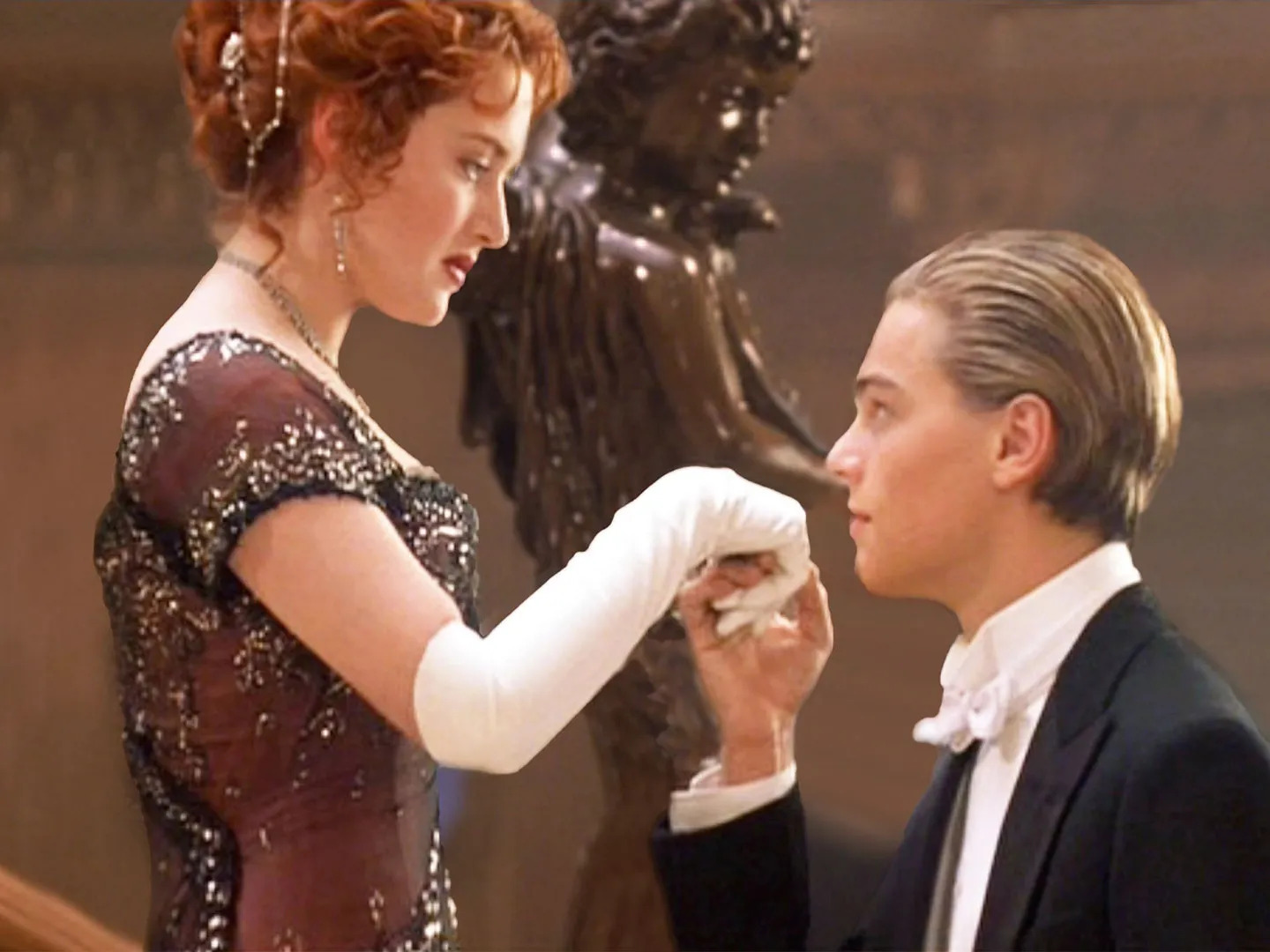 Cena do filme Titanic (1997), em que Jack beija a mão de Rose durante um jantar de gala. Ele veste traje white tie, com casaca preta e gravata-borboleta branca, enquanto ela usa um vestido bordado e luvas longas. | Foto: CBS Arquivo de Fotos